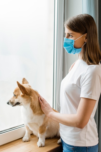 Vista laterale della donna con la mascherina medica che osserva attraverso la finestra con il suo cane