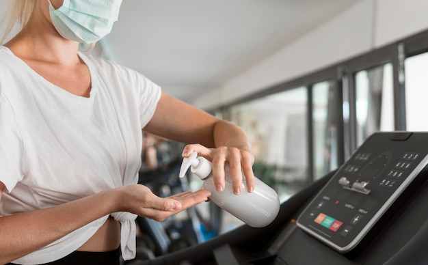 Foto vista laterale della donna con maschera medica utilizzando disinfettante per le mani in palestra mentre sul tapis roulant