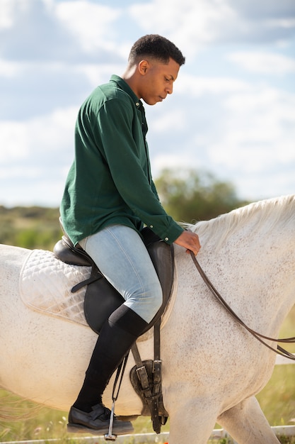 Side view of young African American male looking away while sitting on back of white horse on ranch