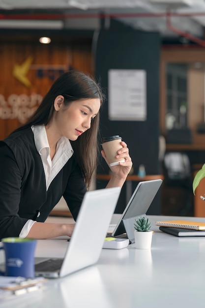 Side view of young asian businesswoman in black suit holding cup of coffee and working on laptop computer while sitting at her office desk.