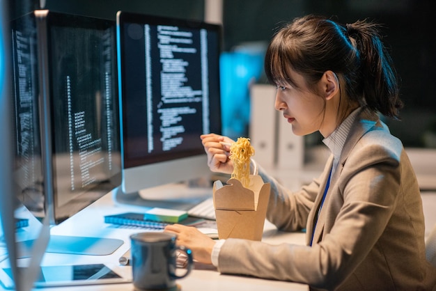 side view of young asian female programmer eating chinese wok during work over coded data in front of computer monitor