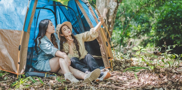 Side view Young Asian pretty woman and her girlfriend sitting at front of tent use mobile phone take photo during camping in forest with happiness together