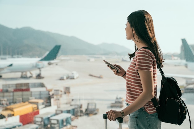 フライトを待っている空港の窓の近くに携帯電話を持って立っている若いアジア旅行女性の側面図。滑走路の飛行機を見渡すラウンジ エリアのホールでバックパックと荷物を持つ美しい少女