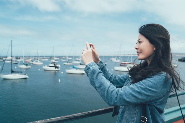 올드 피셔맨스 워프 몬테레이(Old Fisherman's Wharf Monterey)의 앞 좌석에 스마트 폰이 달린 요트 사진을 찍는 젊은 아시아 여성 관광객의 측면. 보트 근처 해변 베이에 핸드폰을 들고 웃는 여성 여행자