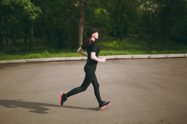 Side view Young athletic brunette girl in black uniform and cap training, doing sport exercises and running, looking straight on path in city park outdoors