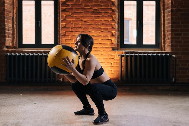 トレーニング中に重いメディシンボールでスクワットをしているスポーツウェアを身に着けている強い体を持つ若い運動女性の側面図