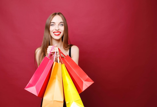 Side view of young attractive smiling woman with bright lipstick in black dress standing in studio with paper shopping bags and looking at camera on red background black friday concept