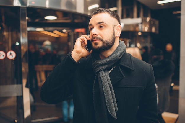 Вид сбоку молодого бородатого мужчины, одетого в обычную одежду, он стоит на улице и использует смартфон