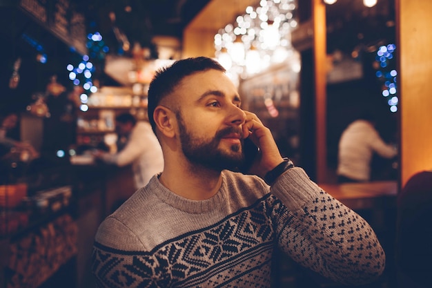 La vista laterale di giovane uomo barbuto, abbigliamento incasuale vestito, sedendosi alla tavola di legno rotonda in caffè con l'interno moderno e sta tenendo lo smartphone.