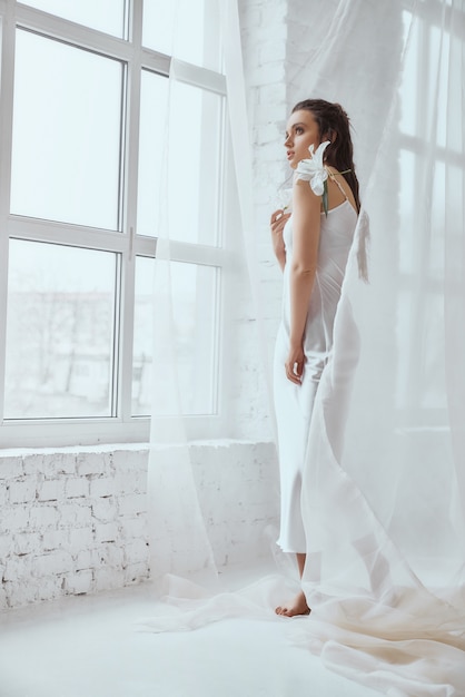 Side view of young beautiful brunette woman standing and holding lily flower. Portrait of girl with wet hair posing on white background and looking out window between tulle. Concept of beauty.