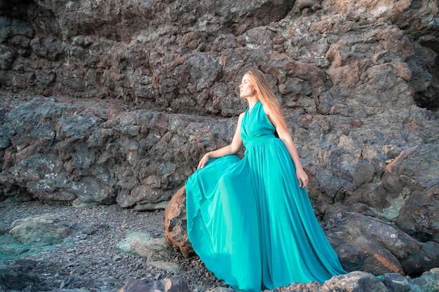 Side view a young beautiful sensual woman in a mint long dress posing on a rock high above the sea