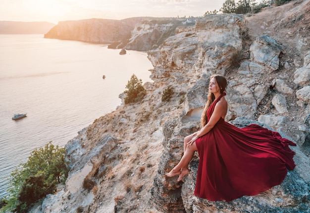 Side view a young beautiful sensual woman in a red long dress posing on a rock high above the sea