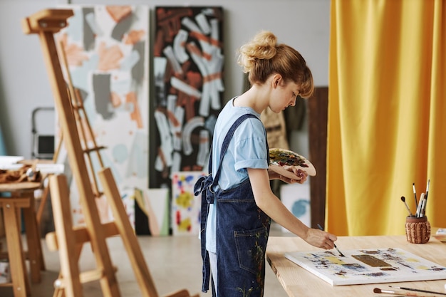 Side view of young blond woman in denim apron painting artwork with acrylic paints while bending ove