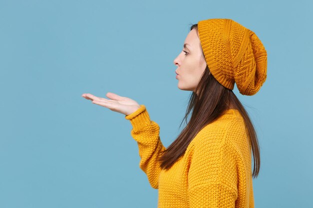 Side view of young brunette woman girl in yellow sweater hat posing isolated on blue background studio portrait. People sincere emotions lifestyle concept. Mock up copy space. Blowing send air kiss.