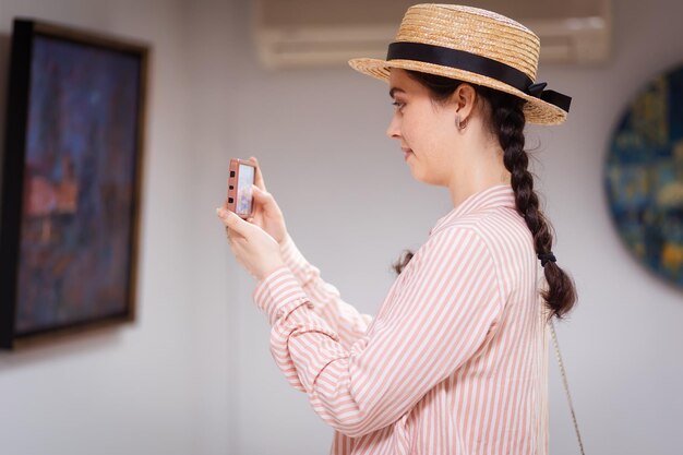 Side view of young Caucasian woman wearing straw hat and takes photo of paintings using smartphone Concept of cultural education and visit museum