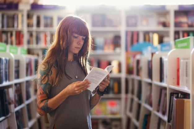 문신을 한 젊은 백인 여성이 서점에서 책을 읽는 포즈를 취하는 측면 시각 교육과 쇼핑의 개념