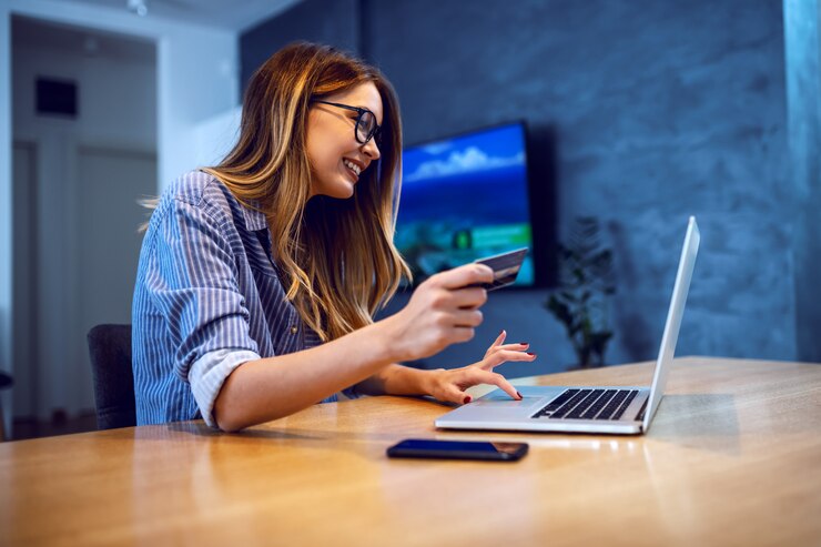 Photo side view of young charming caucasian woman with eyeglasses holding credit card and using laptop for online shopping while sitting at dining table at home.