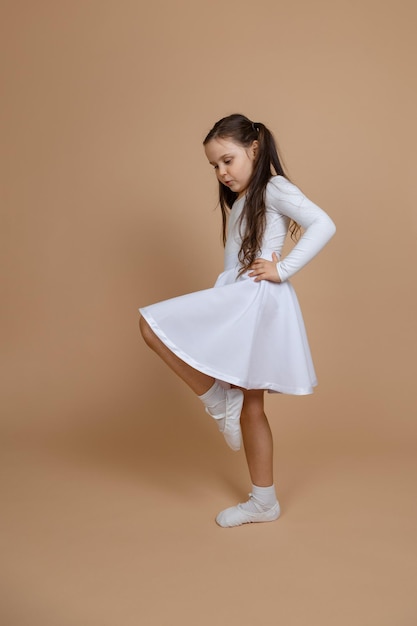 Side view of young concentrated girl with long dark hair in white dress socks and gymnastics shoes standing holding hands on waist raising left leg up on brown background Ballet ballroom dance