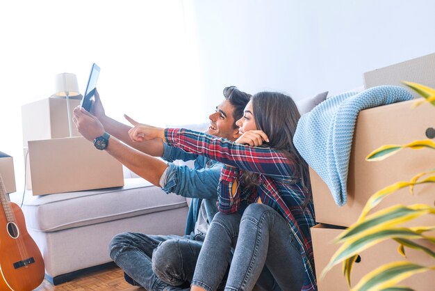 Side view of young couple sitting on floor near carton boxes and browsing tablet while mov