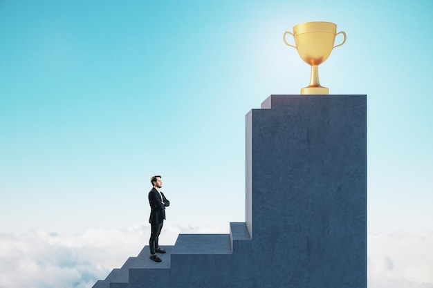 Side view of young european businessman standing on stairs to golden winner's cup with obstacle on blue sky background success and career growth concept