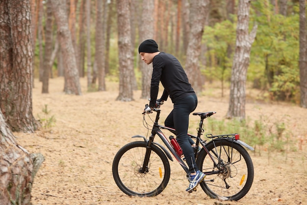 Side view of young European male riding bike in forest on sunny day, sporty man wearing black sportwear and cap onbicycle in wood