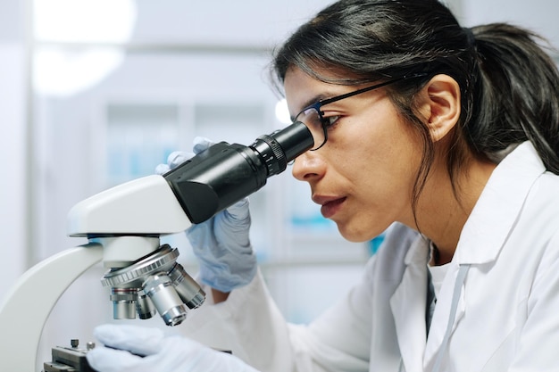 Вид сбоку молодой женщины-биохимика, смотрящей в микроскоп во время исследования