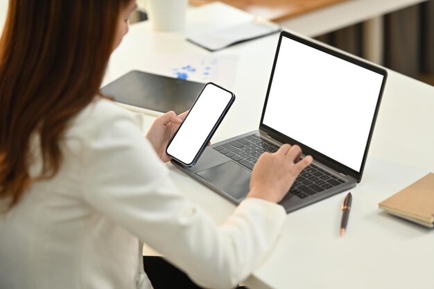 Вид сбоку на молодую сотрудницу, сидящую за рабочим столом и печатающую вручную на ноутбуке и использующую смартфон