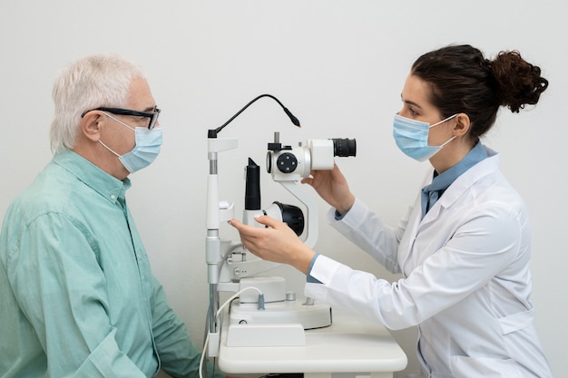 Vista laterale della giovane oculista femminile che utilizza attrezzature mediche nelle cliniche durante il test della vista del paziente maschio anziano in maschera protettiva