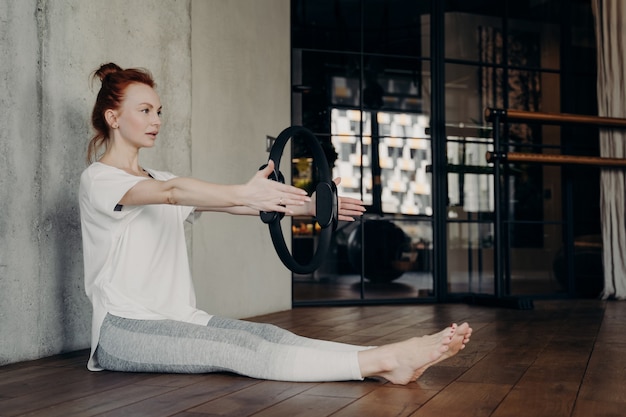 Side view of young focused sportive female sitting on floor with outstretched legs in fitness studio and doing arm exercising with pilates ring, wearing white fitness t-shirt and grey leggings