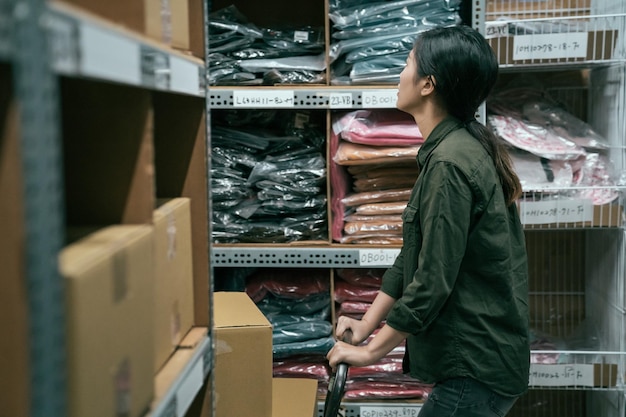 Side view young girl worker searching products in warehouse of\
company. woman staff pushing cart walking in stockroom. female\
employee wearing uniform prepare parcel for customer in\
storehouse.