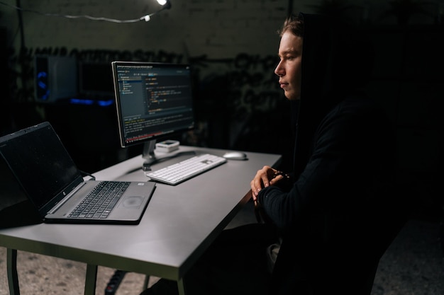 Side view of young hacker man wearing longsleeved shirt\
stealing information preparing to break password sitting at desk in\
dark room