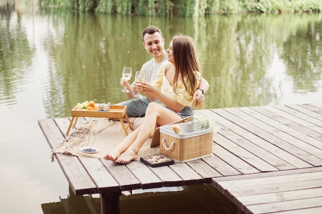 川や湖の近くのロマンチックなピクニックで若い幸せなカップルの側面図、一緒に屋外でワインを飲む女性と男性、夏休みを楽しんでいる人々、ライフスタイルの写真