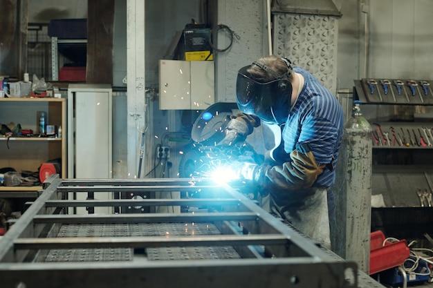 Side view of young male welder in protective face mask carrying out repair work