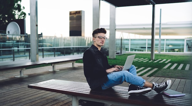 Side view of young man in casual clothes and glasses typing on laptop and sitting on wooden bench