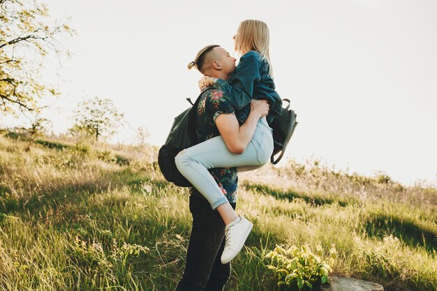 Side view of young man in casual summer clothes holding in arms happy attractive blond woman hugging him on bright green sunlit grassy hill