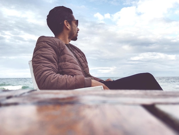 Side view of young man sitting on beach against sky