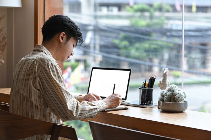 Photo 측면 보기 젊은 남자는 카페에서 노트북 컴퓨터 앞에 앉아서 노트북에 메모를 합니다.