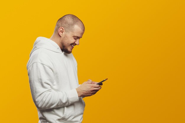 Side view of young man wearing white hoodie using mobile cell phone isolated over yellow background