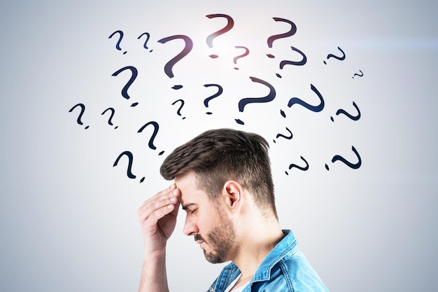 Side view of young man with beard wearing jeans shirt and having strong headache standing near white wall with question marks drawn on it. Concept of difficult problem