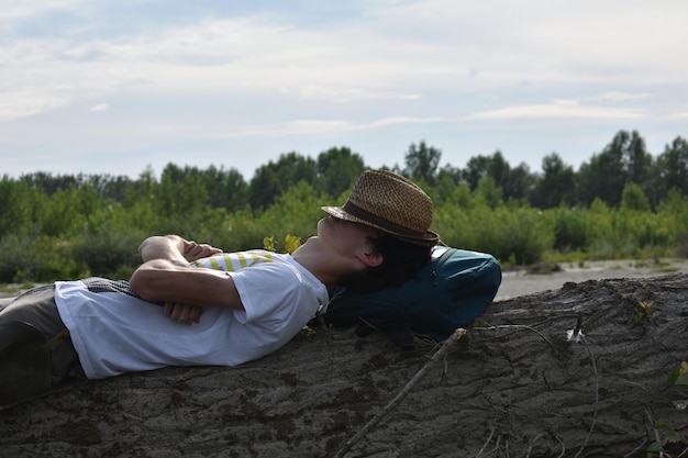 Photo side view of young man with hat and backpack lying on tree trunk against cloudy sky