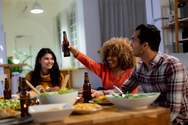 若い女性がビールのボトルを持ち上げて乾杯し若い男性と若い白人の女性と一緒に感謝祭の夕食に用意されたテーブルに座っています