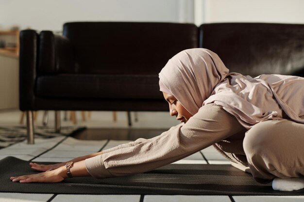 Side view of young Muslim woman stretching her arms forwards while sitting on mat