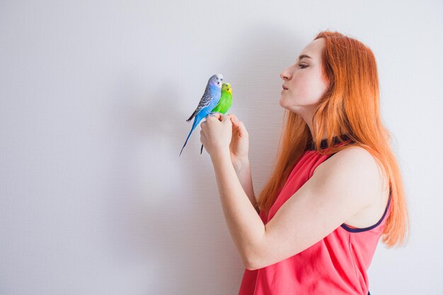 Side view of a young redhaired woman is standing against a white wall and holding two beautiful parrots