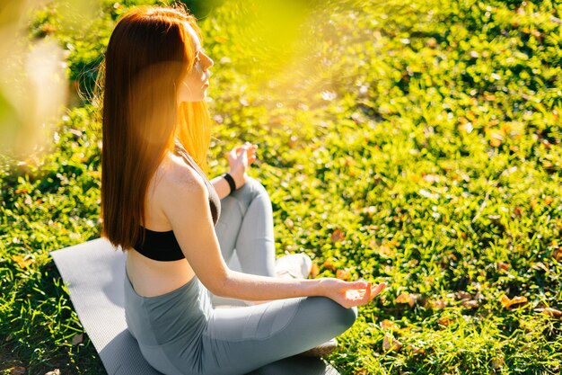 晴れた夏の朝に都市公園で緑の芝生の上のヨガマットに座って膝の上で手をつないで蓮のポーズで瞑想する目を閉じて若い赤毛の女性の側面図
