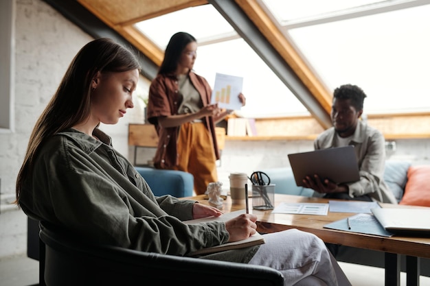 Side view of young serious businesswoman with pen and notebook making notes