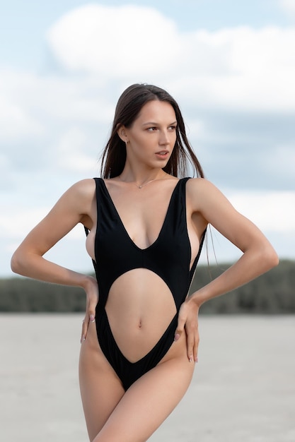 Side view of young sexy female in bikini standing on seashore on sunny day and looking at camera