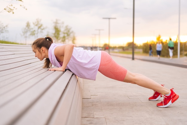 Vista laterale di una giovane donna sportiva che indossa un allenamento activewear facendo flessioni usando una panchina da strada in