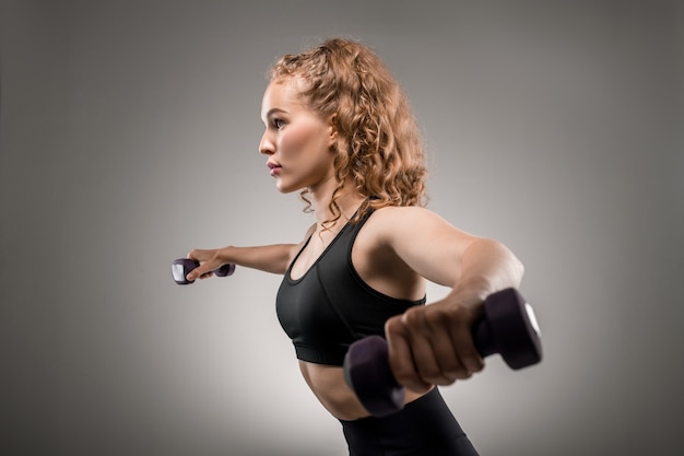Side view of young sportswoman in black tracksuit lifting dumbbells and breathing during physical exercise on grey