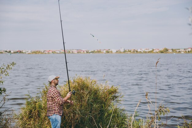 Side view Young unshaven man with fishing rod in checkered shirt, cap and sunglasses casts fishing pole on lake from the shore near shrubs and reeds. Lifestyle, recreation, fisherman leisure concept.