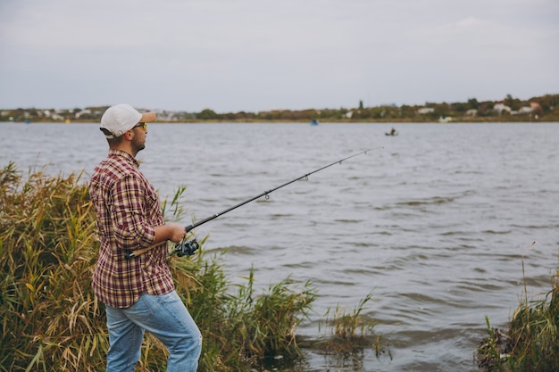 Vista laterale giovane uomo con la barba lunga con una canna da pesca in camicia a scacchi, cappello e occhiali da sole lancia la canna da pesca su un lago dalla riva vicino a arbusti e canne. stile di vita, ricreazione, concetto di svago del pescatore